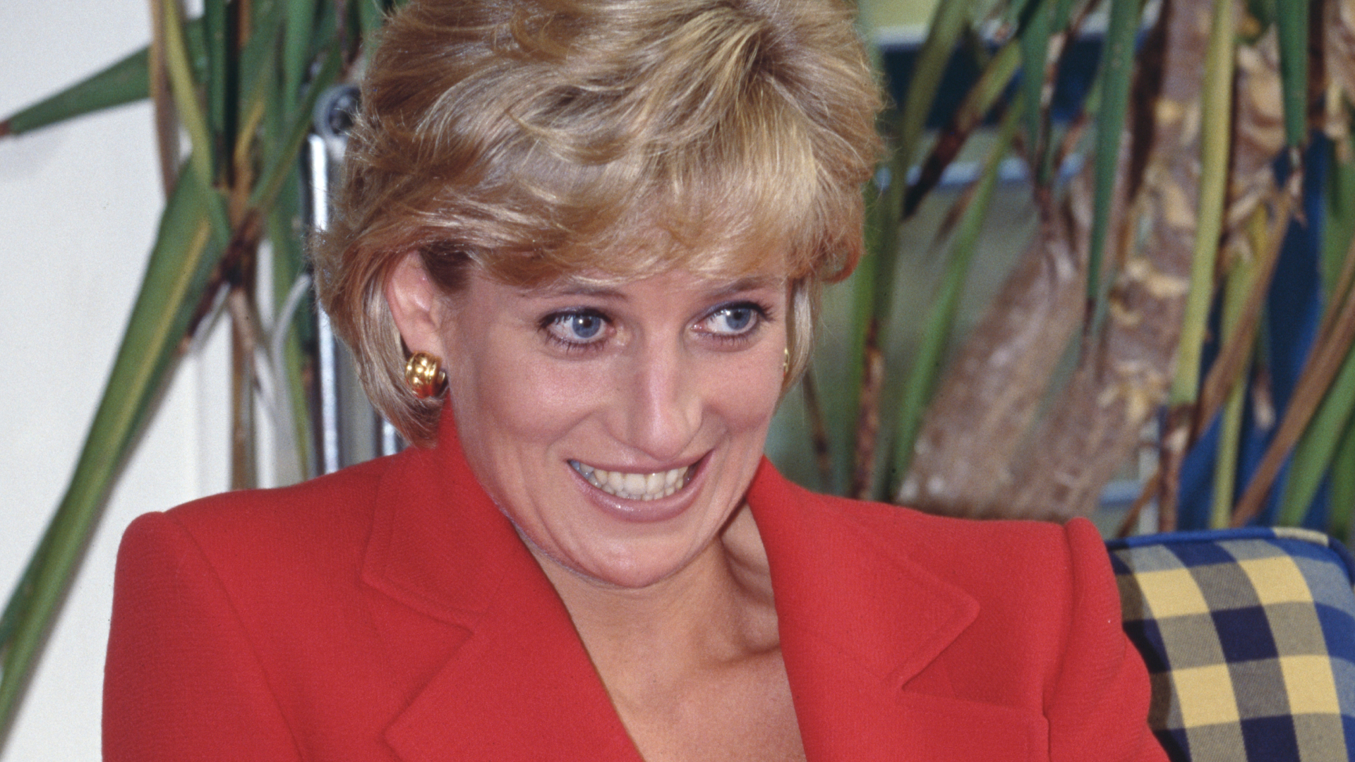 Princess Diana has short blonde coiffed hair and wears a bright red jacket with a black top underneath and looks sheepish