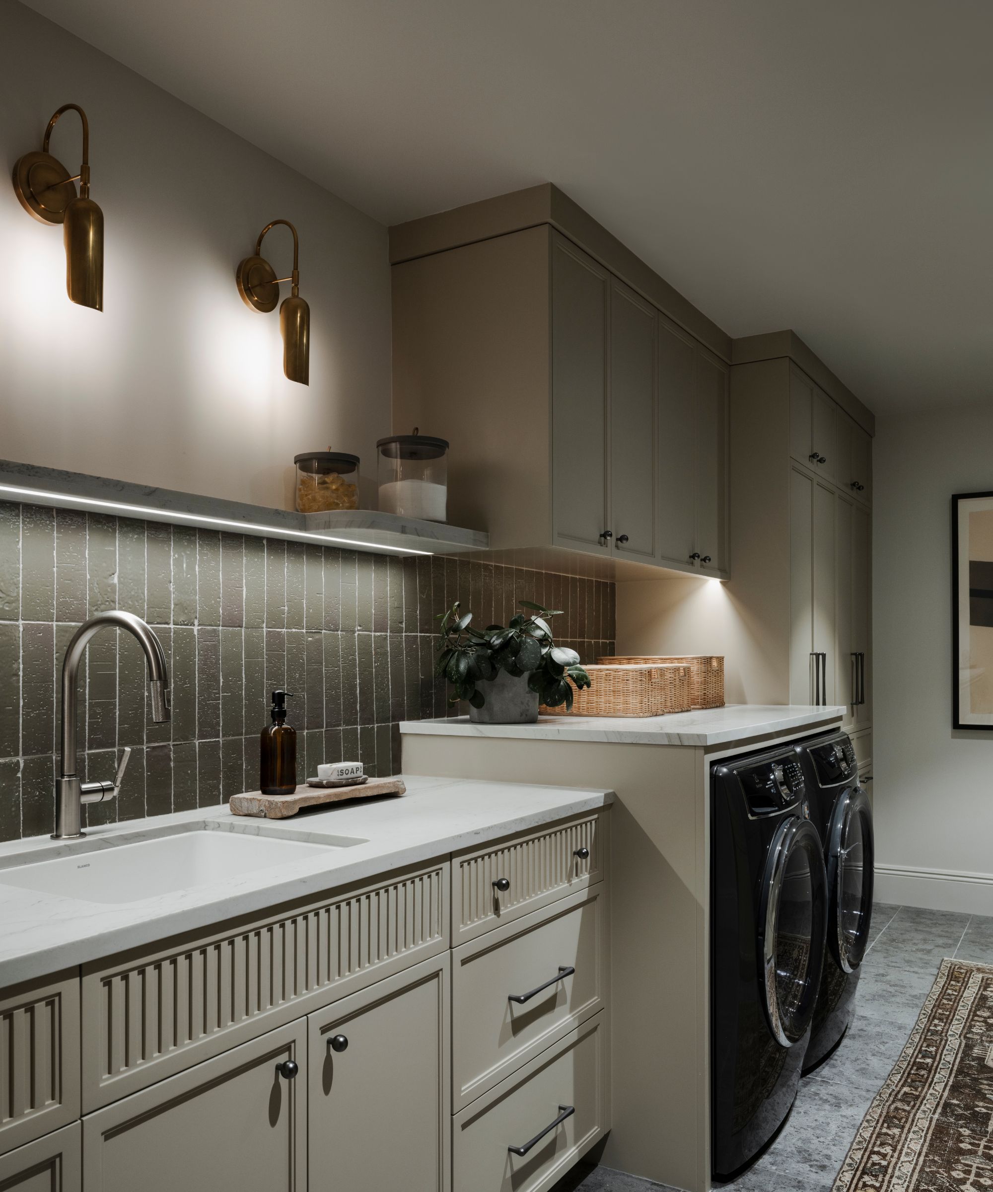 A laundry room with taupe cabinets, neutral walls, two wall sconces, and green vertical backsplash tiles.
