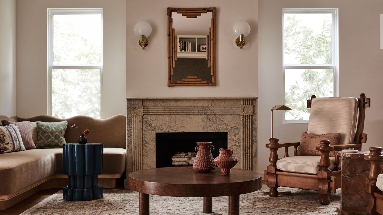 A light pink living room with a dusty rose and warm brown color scheme. There is a tan marble fireplace in the middle with a bamboo mirror over it. There is a brown velvet corner sofa with scalloped edges and a dark wooden coffee table, and accent chairs. 