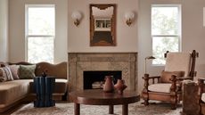 A light pink living room with a dusty rose and warm brown color scheme. There is a tan marble fireplace in the middle with a bamboo mirror over it. There is a brown velvet corner sofa with scalloped edges and a dark wooden coffee table, and accent chairs. 