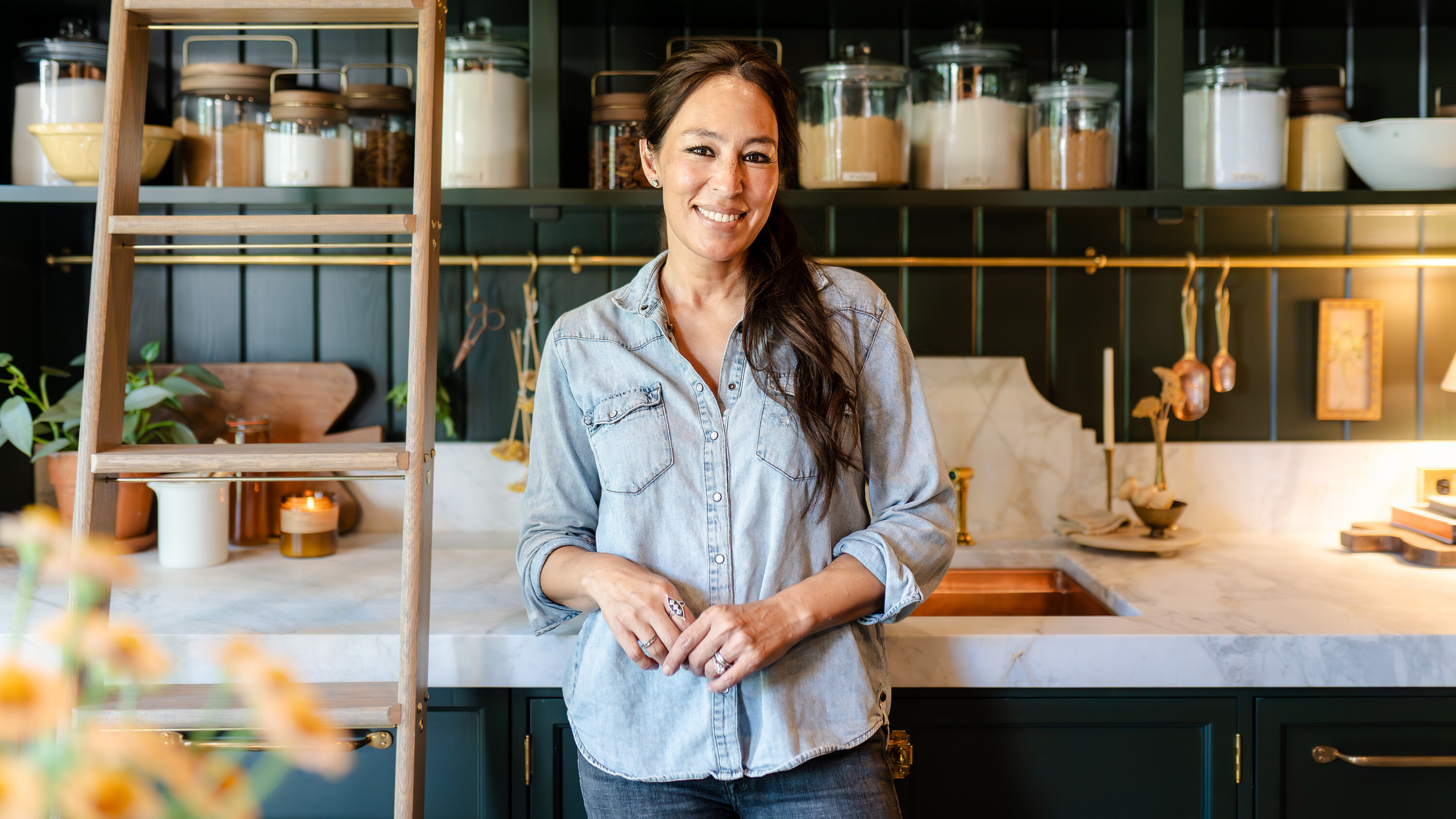 Joanna Gaines standing in a kitchen with jars of grains, green painted cabinets, and white marble countertops