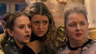 Jen (Máiréad Tyers), Carrie (Sofia Oxenham) and Mary (Siobhán McSweeney) wearing party gear huddle together looking at something that has shocked them in Extraordinary.