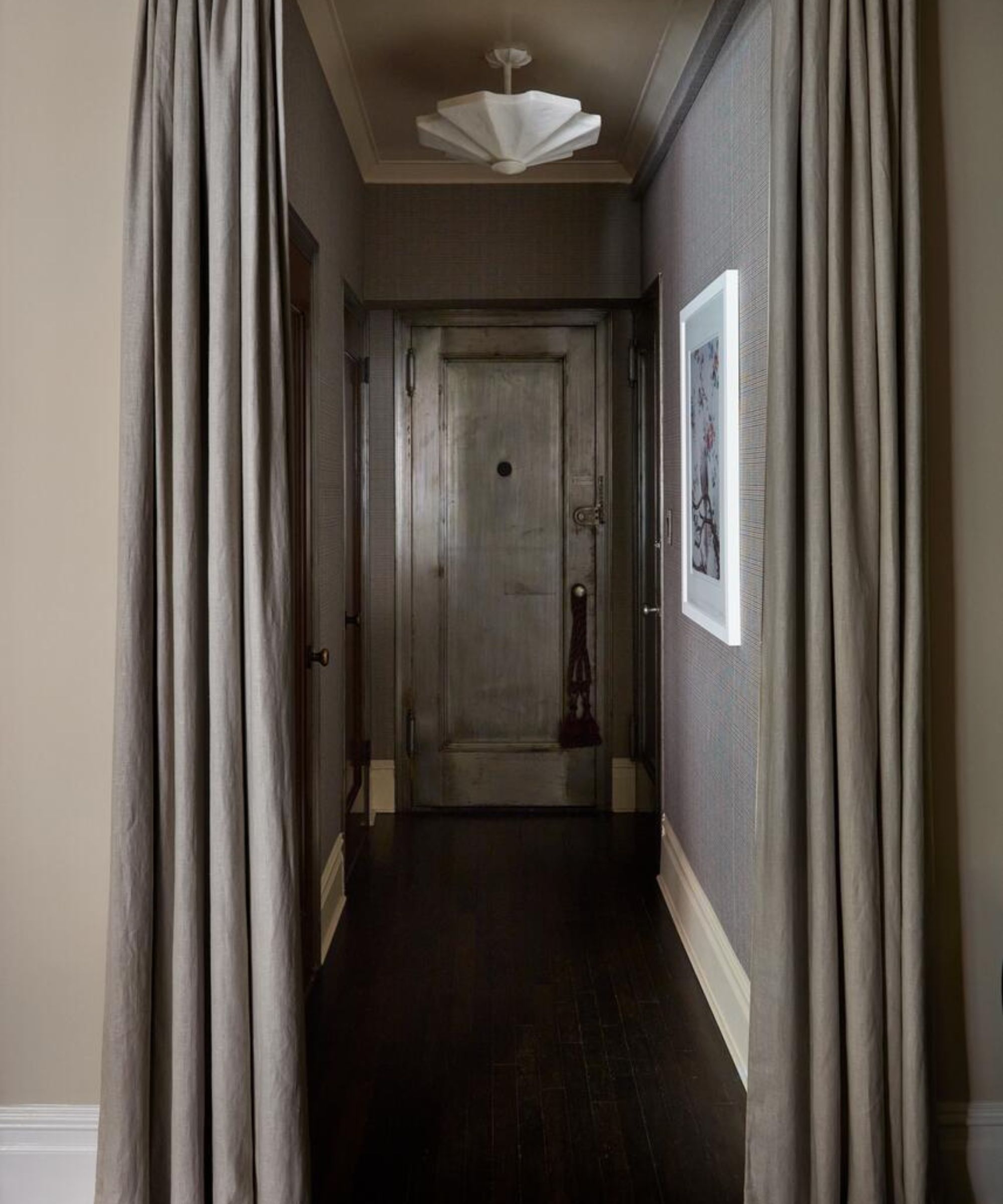 A symmetrical hallway with dark wood floors and grey textured wallpaper, framed by heavy floor-to-ceiling drapes and leading to a metallic finished door