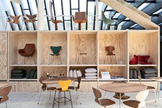 Inside pavillion display of Fritz Hansen’s anniversary collection, wooden display unit, chaoirs, tables, cushions on display, neutral floor, wooden and glazed slanted roof frame in the backdrop