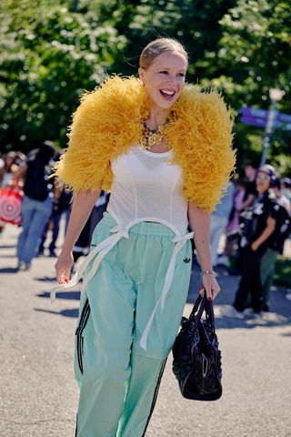 Chloe King at New York Fashion Week Spring 2025 wearing blue Adidas track pants, a white bow tank top, yellow feather shrug, gold charm necklace, and black handbag.