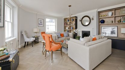 Grey living room with 2 orange chairs and 2 grey couches around a fireplace with brass decorations in the room