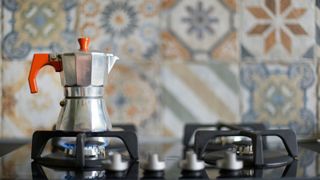 A stainless steel moka pot on the stove with a tiled wall behind it