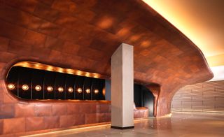 Copper curved wall with light fixtures and a wide column supporting the top part. Dark wooden boards cover the other wall and brown tiles are on the floor in the Sea Containers hotel.