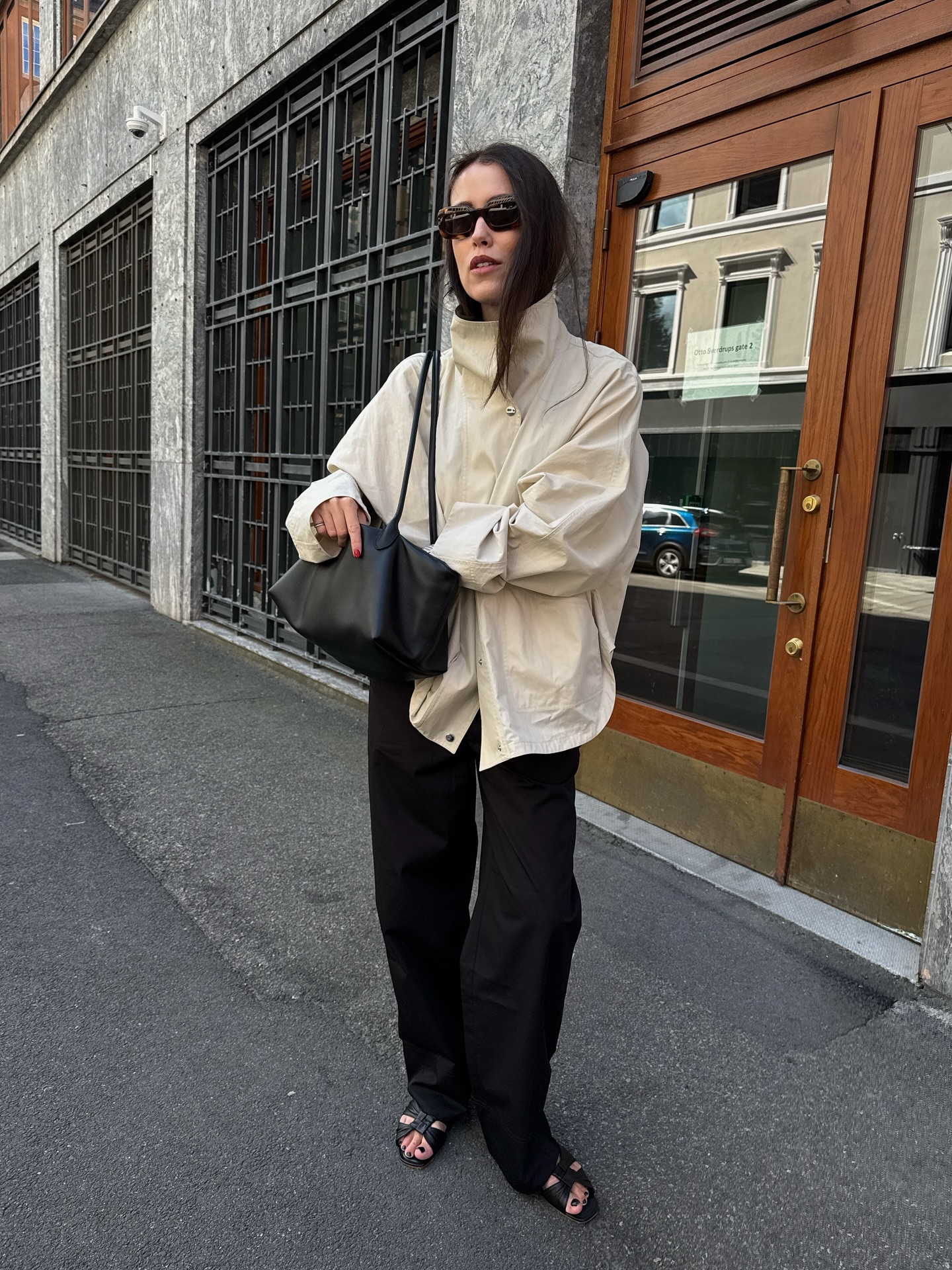 An image of an influencer wearing a beige funnel-neck jacket, black satin trousers and black sandals whilst carrying a black tote