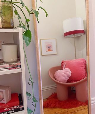 A pink mirror reflecting a pink chair with a lamp over it, next to a white shelf
