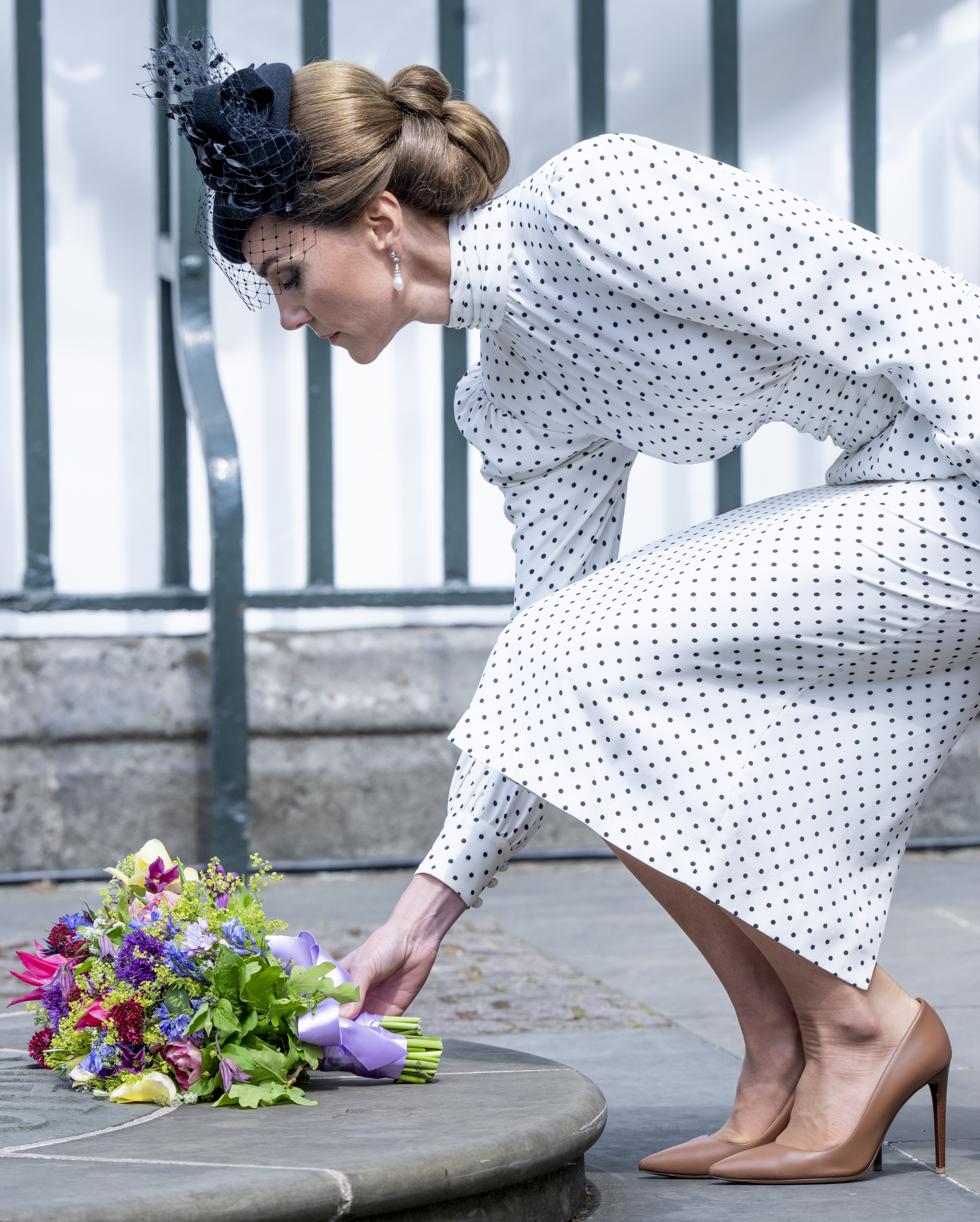 Princess Kate wearing a white polka dot dress kneeling down to place flowers on the ground