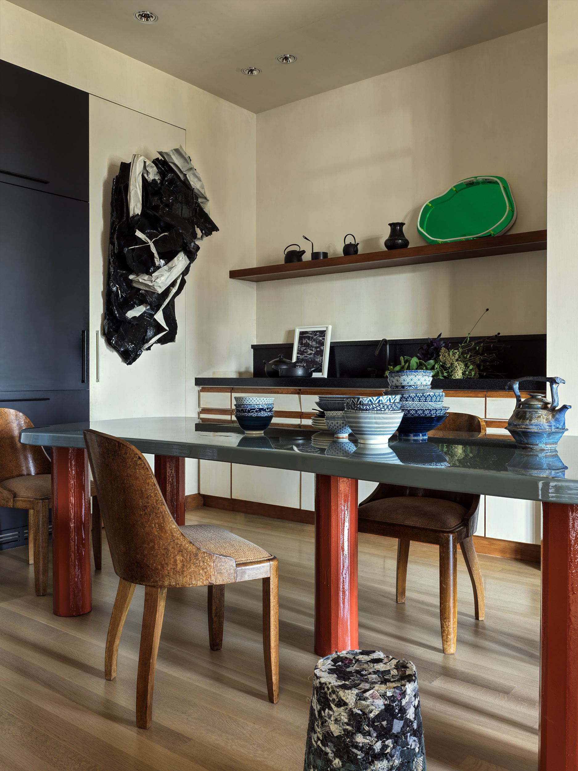 A kitchen with a large central island table with a blue top and red legs, with wood chairs round it and a large black and white artwork sculpture on the wall next to the wood open shelf