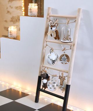 A mini light wooden ladder with dip paint effect, dressed with baubles and reindeer decor leant against stairs and on checkered monochrome floor