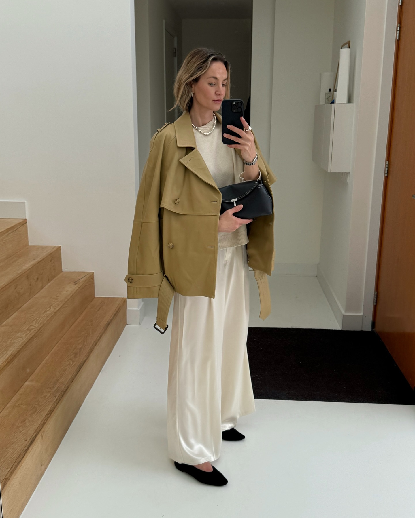 An image of an influencer wearing a cropped beige trench coat, a cream knit, cream satin trousers and black ballet flats