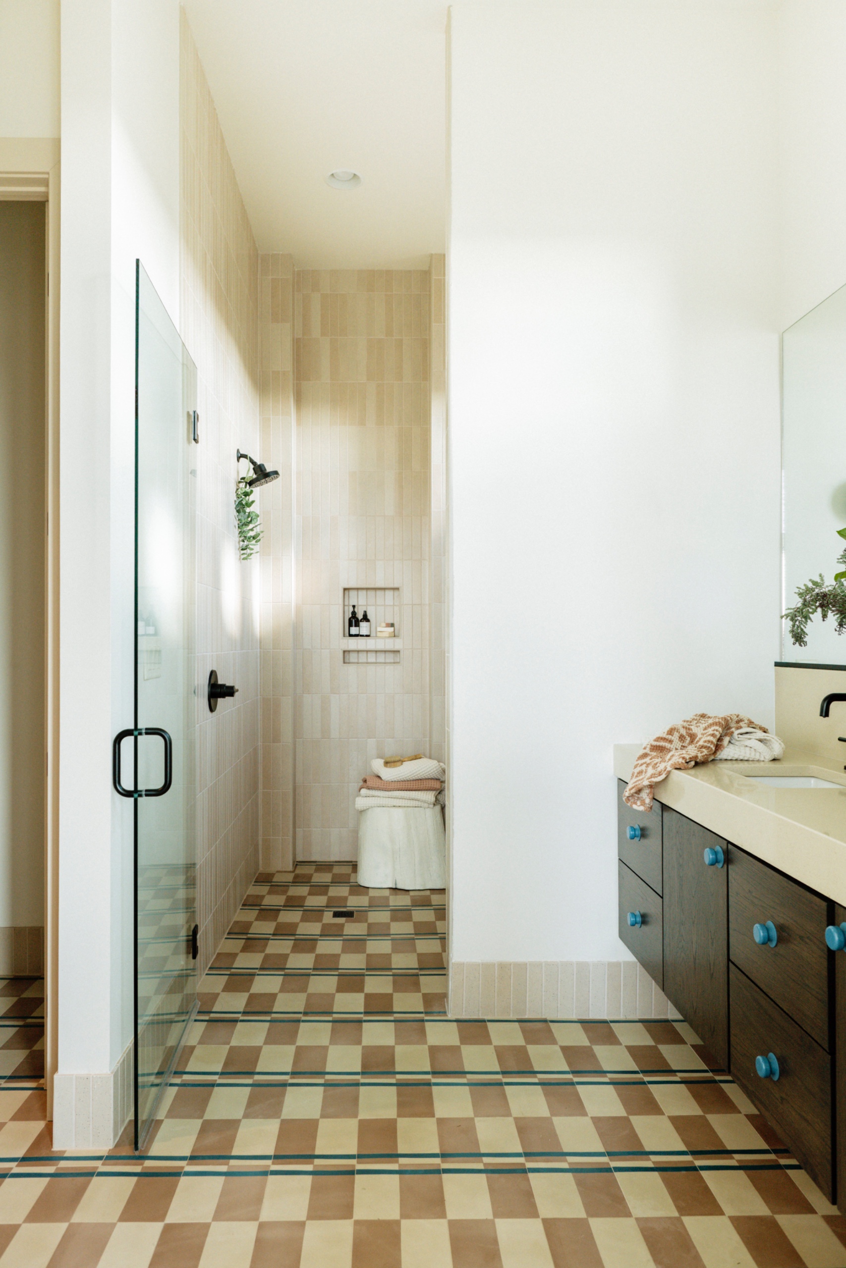 Glass shower enclosure with caramel and cream checked floor tiles crossed by blue stripes, pale vertical wall tiles and a wood vanity with blue knobs.