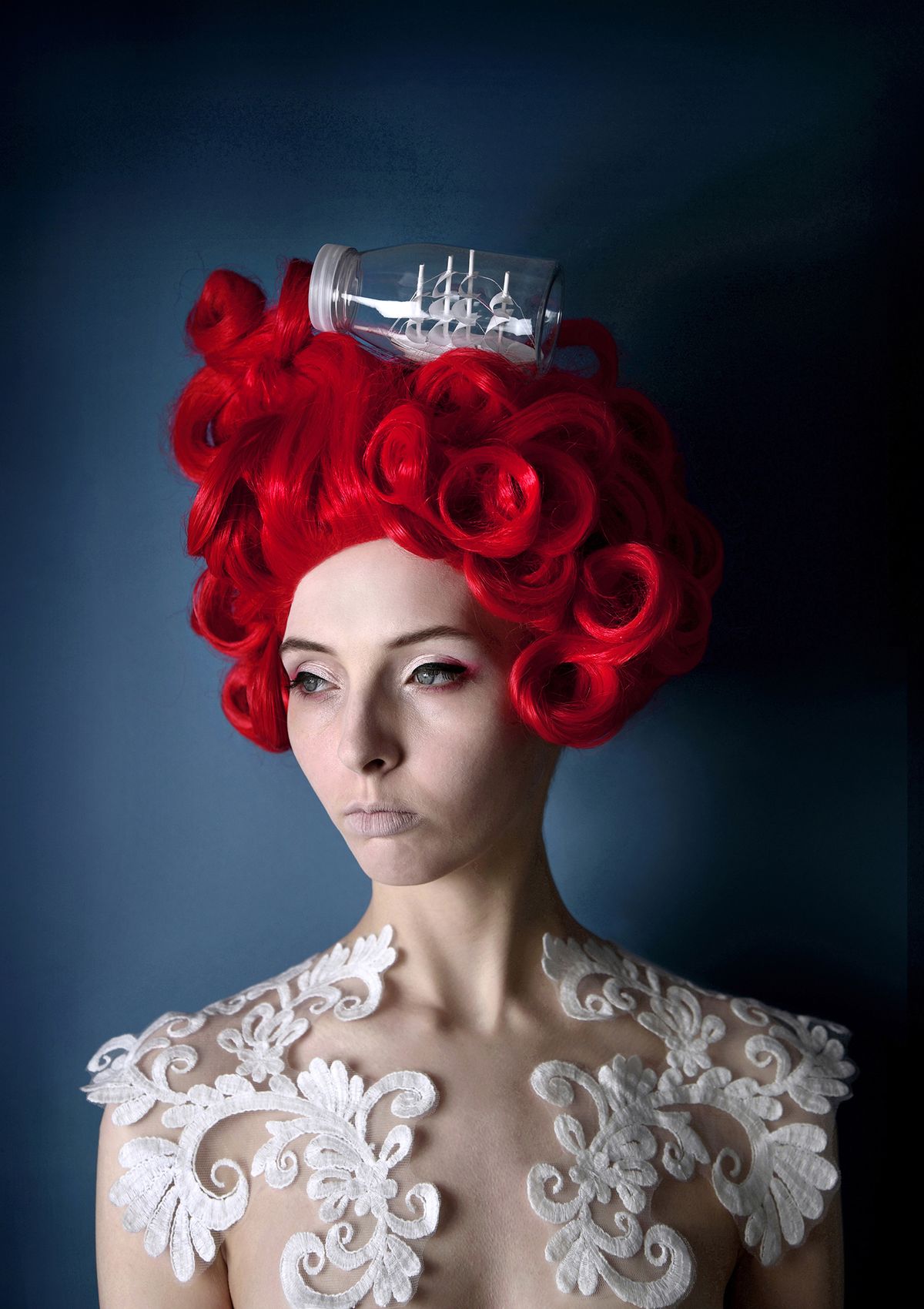 A woman with vibrant red curly hair topped with a glass bottle containing a small ship, adorned with intricate white lace