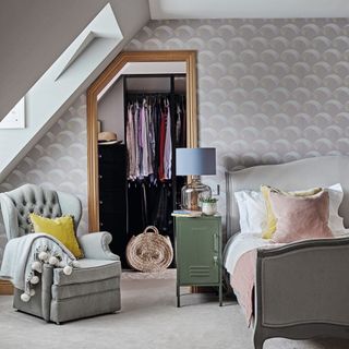 bedroom with sloping eaves, grey patterned wallpaper, armchair and bed with cushions