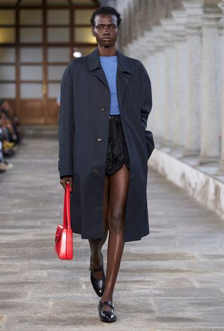 a photo of a model on Bally's spring runway wearing a black bubble skirt with a blue t-shirt and navy trench coat and black mary jane flats