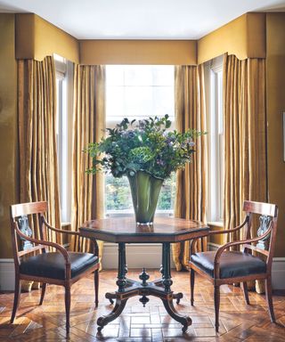 Dining table with two dining chairs with a vase and large flower arrangements, bay window with yellow curtains and pelmet
