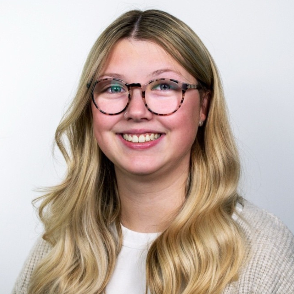 Smiling woman with long blonde wavy hair, tortoiseshell glasses, wearing beige knit against plain grey backdrop.