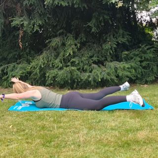 Fitness editor Ruth Gaukrodger performs a Pilates swimming exercise outside on a yoga mat. She is lying down, her body forming an 'X' shape, and her arms and legs are lifted off the ground. Behind her we see a pine tree and grass.