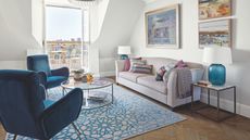 A sunny living room with blue velvet chairs, a white and blue patterned rug, and a pale beige couch with colorful cushions. To the right of the couch is a marble and metal coffee table with a large blue lamp with a white shade, and above are framed painted artworks. In the centre of the room is a circular glass coffee table. Open doors onto a balcony are seen in the background.