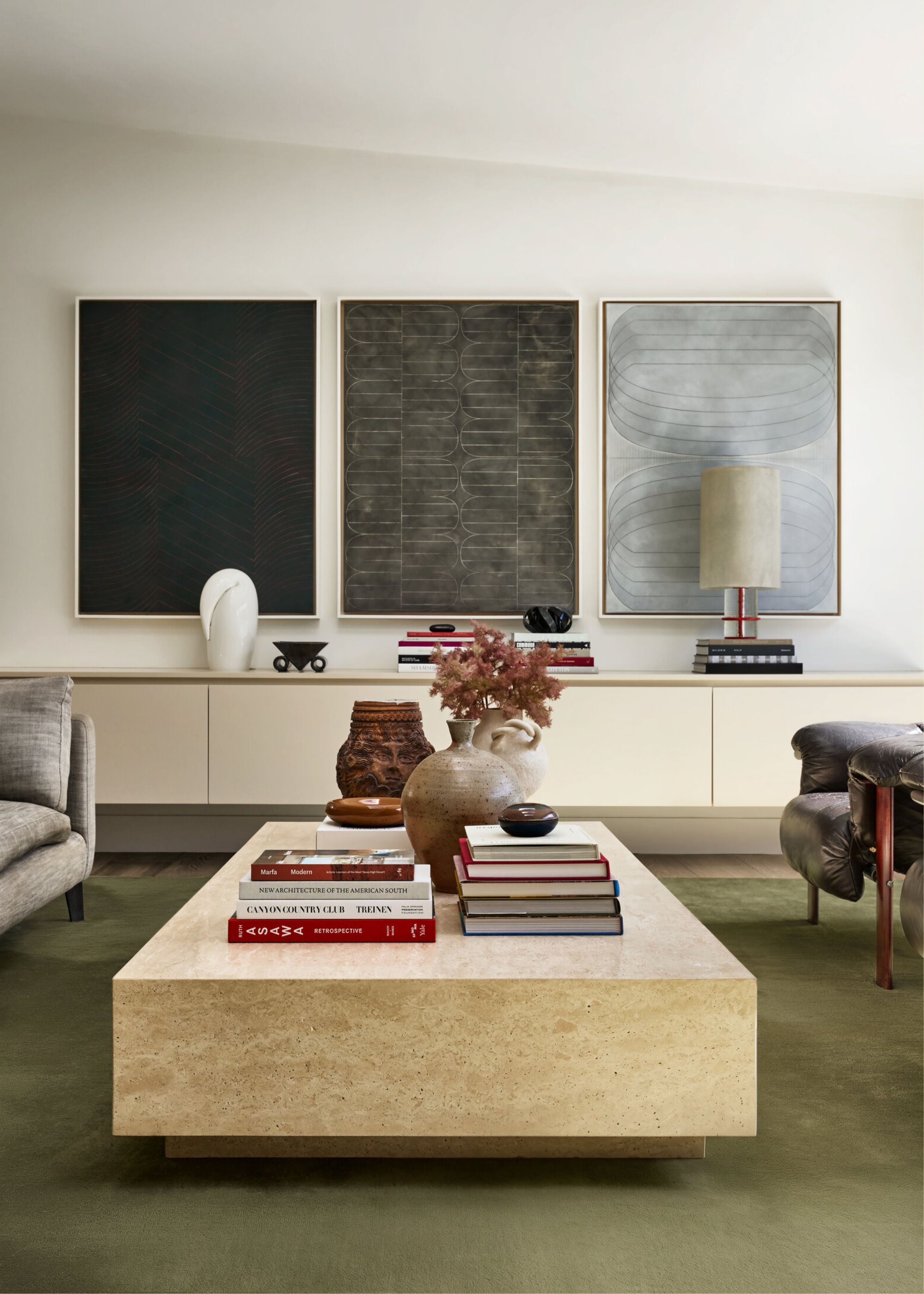 A white living room with a marble coffee table, a stack of books, three paintings, and vases of flowers