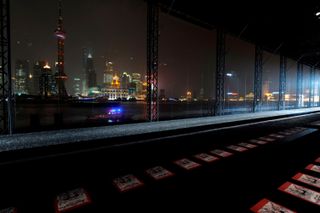 Night time image of city skyline, tall light up buildings in Shanghai, China, 2009, boat on water, red, white and black prints in a line on the floor at the front of the shot
