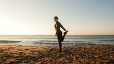 Woman standing on one leg, doing one of the best balance exercises, on beach in activewear against backdrop of the sky and sea