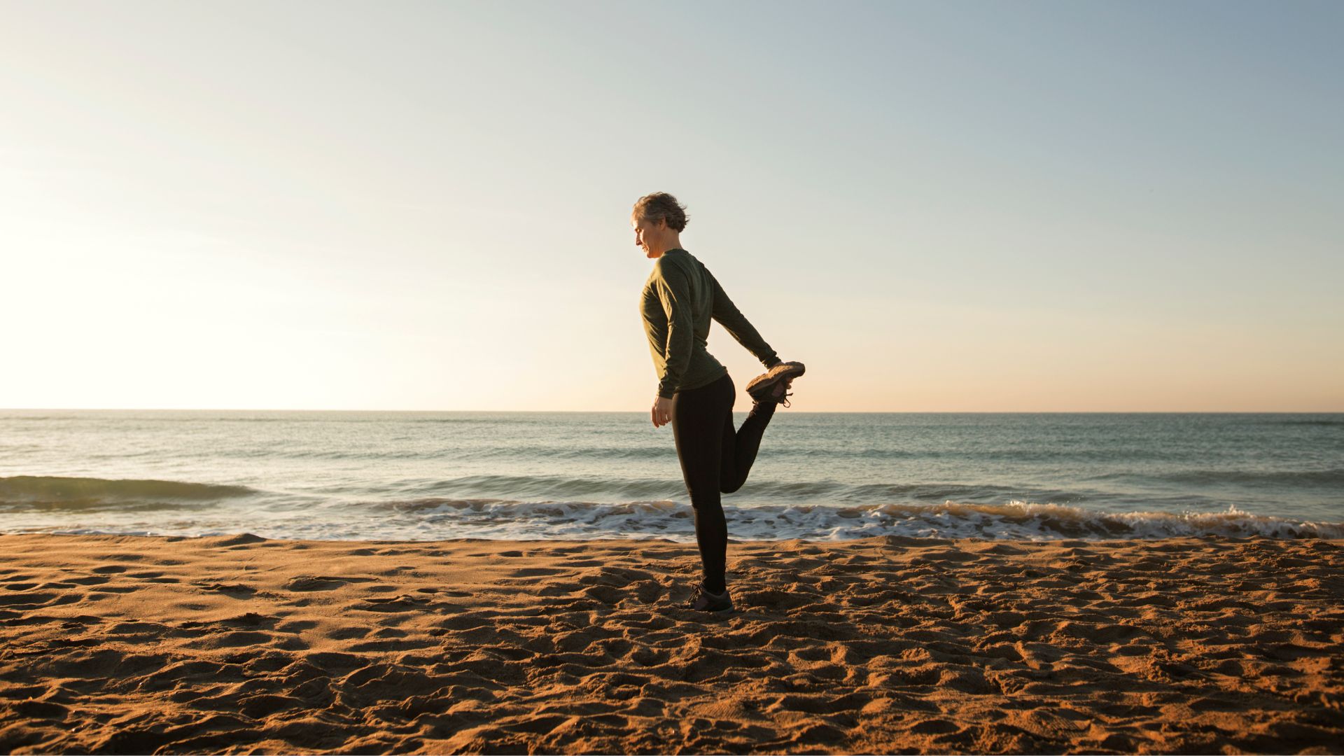 Woman standing on one leg, doing one of the best balance exercises, on beach in activewear against backdrop of the sky and sea