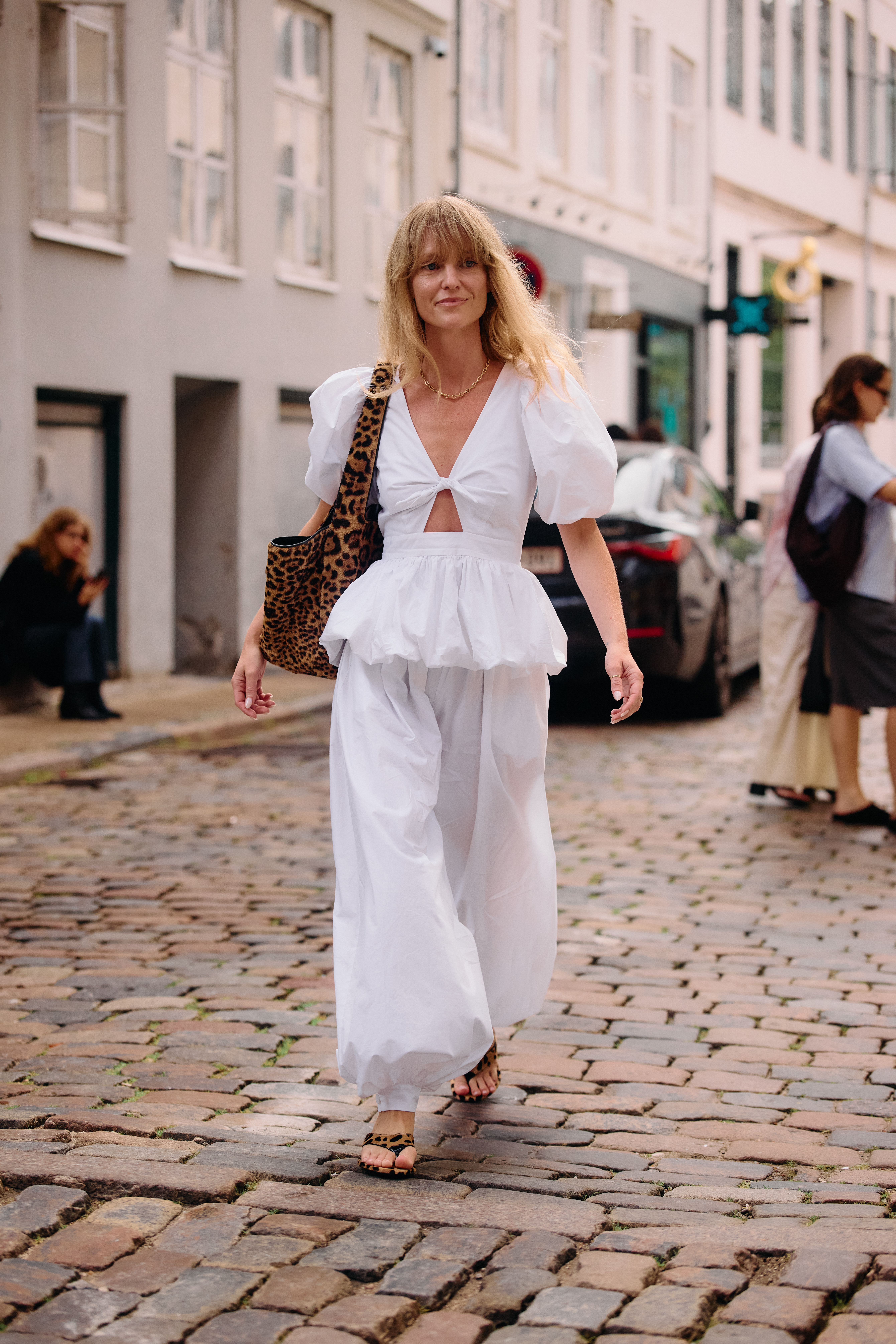 A woman wears a white top, white baggy pants, leopard heels, and a leopard bag.