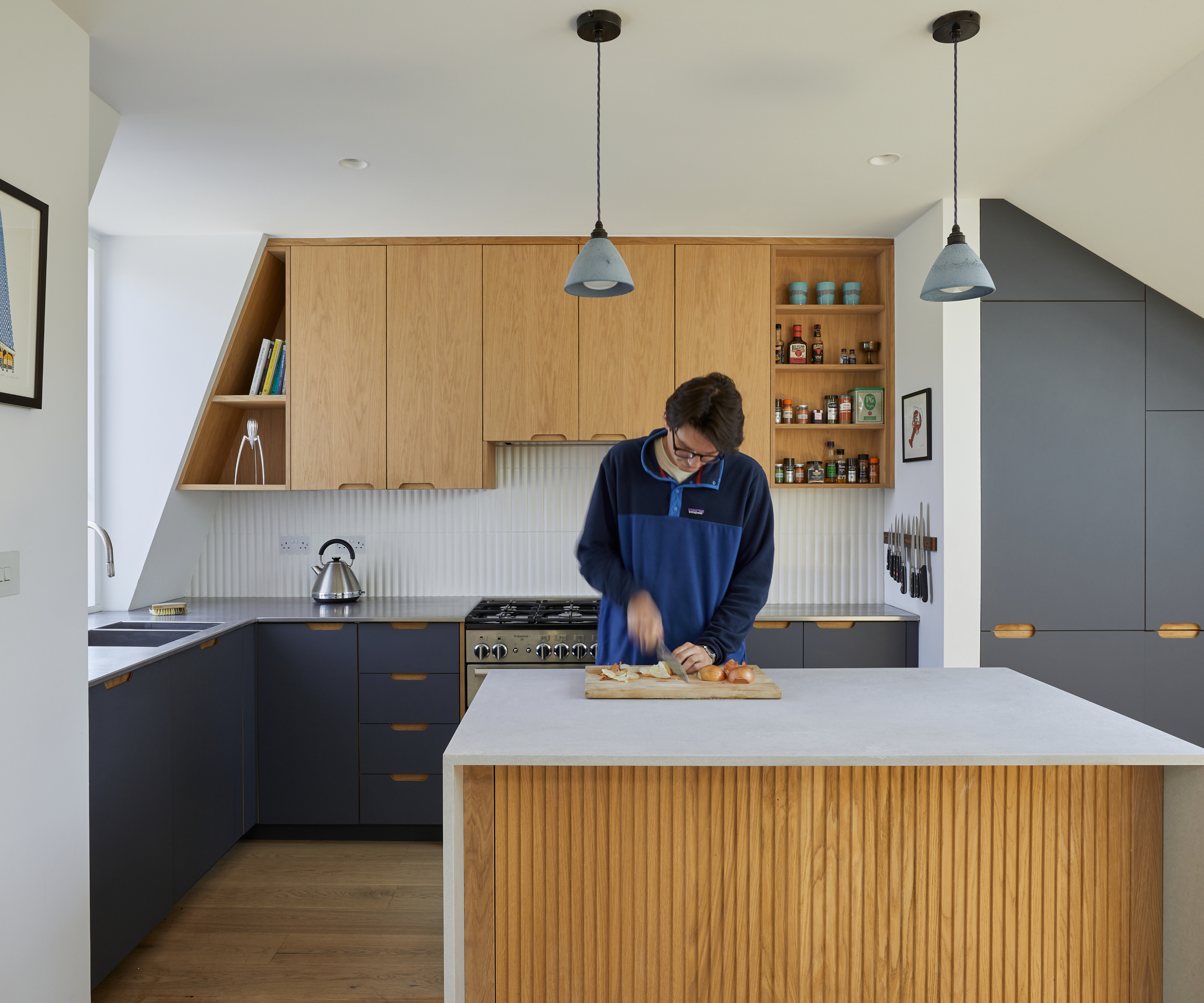 man chopping food at kitchen island in dormer loft conversion featuring white worktops, grey and wooden kitchen cabinets, small pendant lights above island, wooden floor and white walls