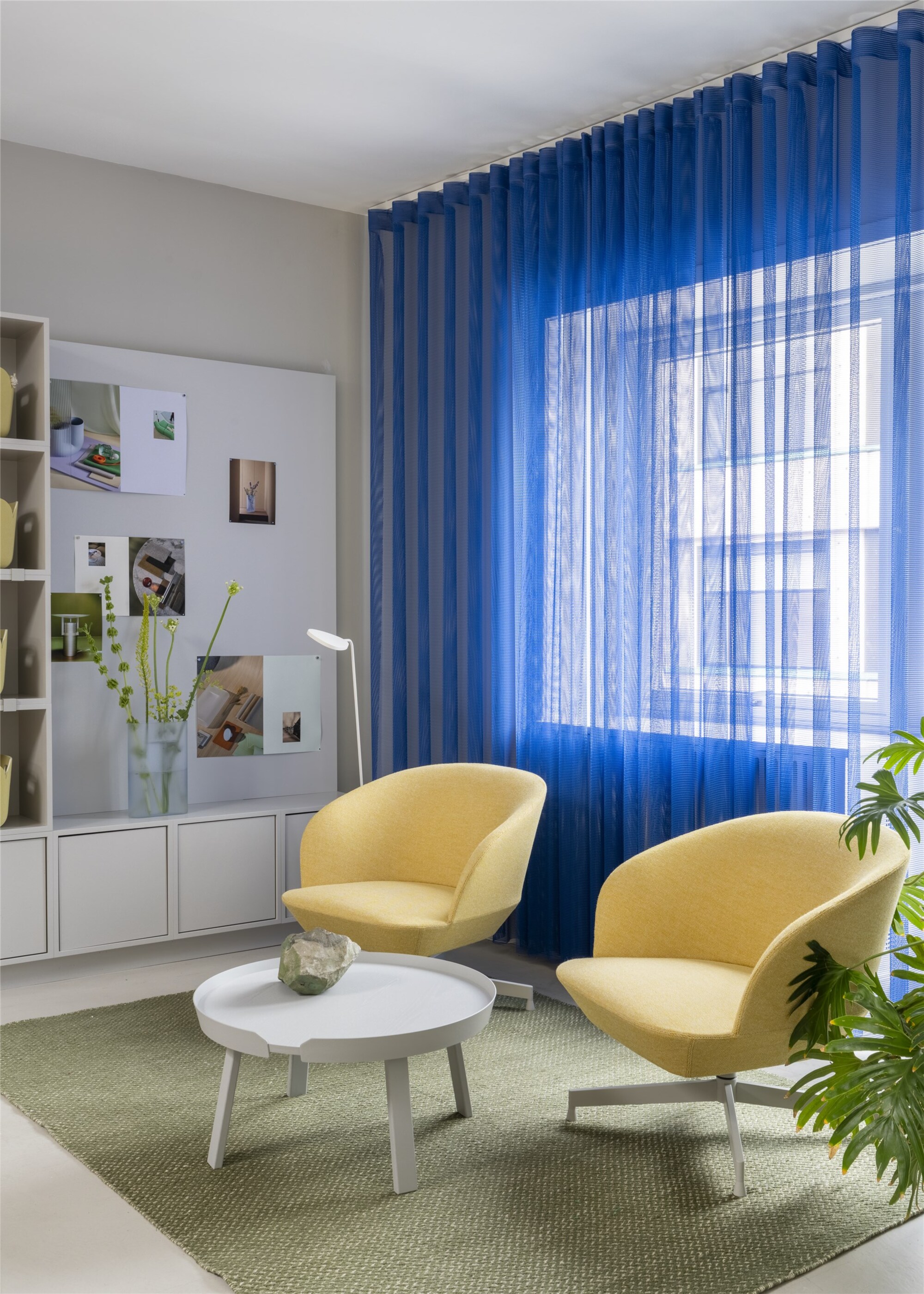 office space with bright blue sheer curtains, pin board in built-in cabinetry, two butter yellow chairs on a green rug with a white coffee table positioned in front