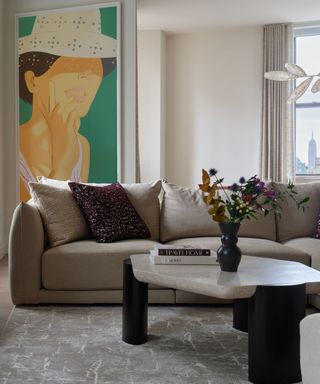 living room with neutral walls, beige sofa, modern sculptural coffee table with vase of flowers, bold green and yellow artwork on the wall