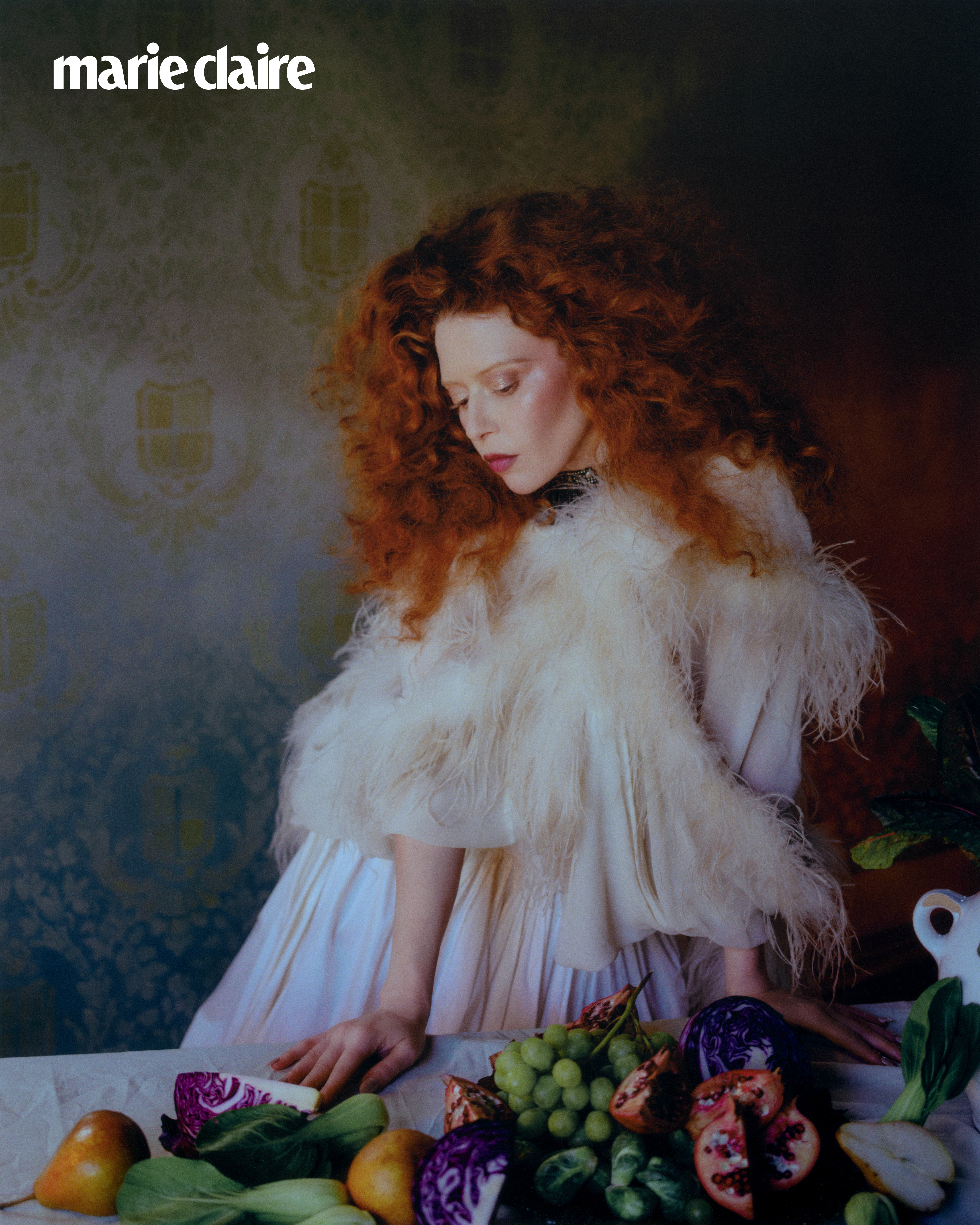 Natasha Lyonne posing by fruit on a table wearing a feathered cape