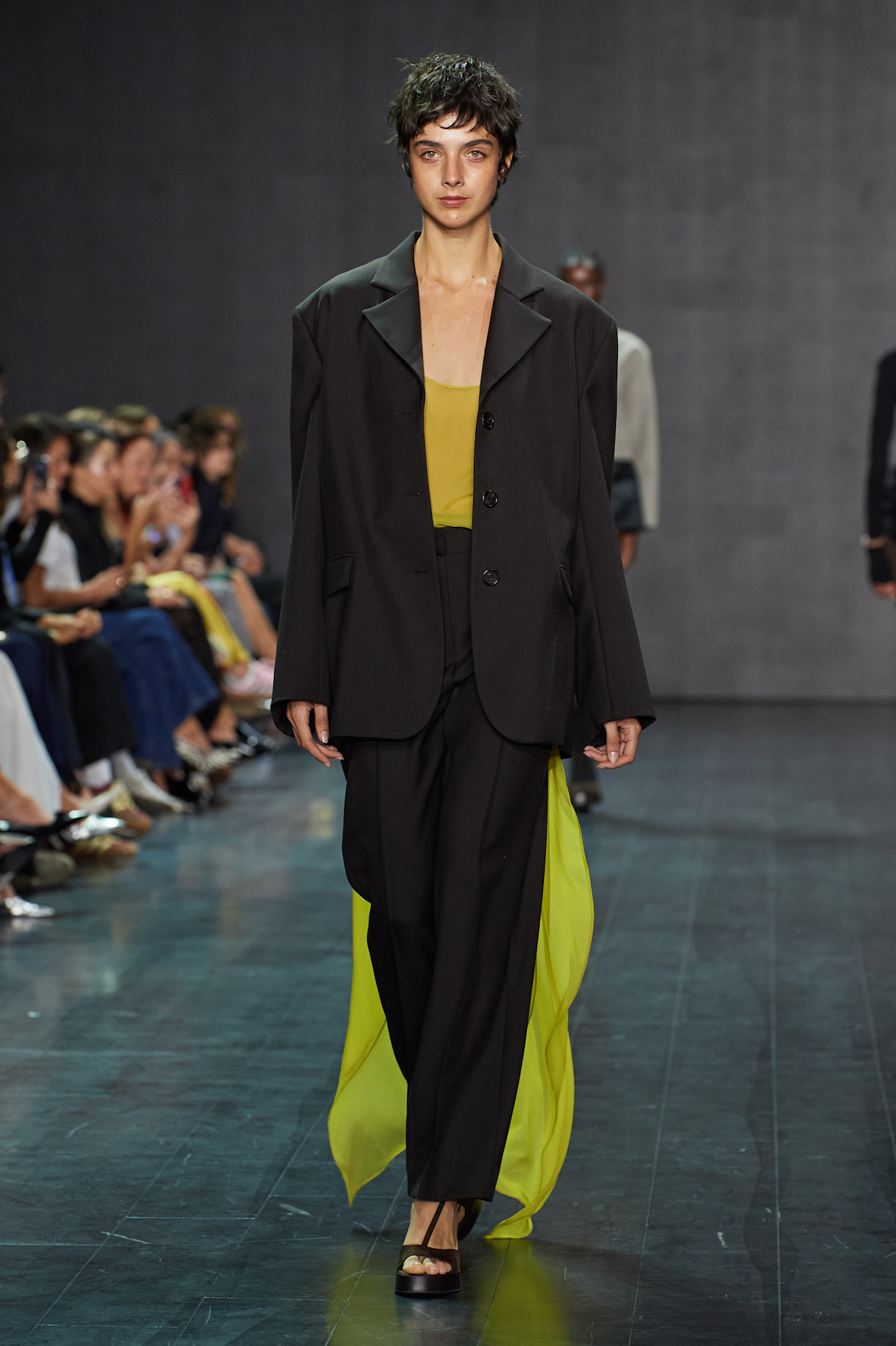 A Beare Park model wearing a black tuxedo blazer layered over a sheer organza slime green tank top that has a train, black wide leg trousers, and black T-strap sandals.