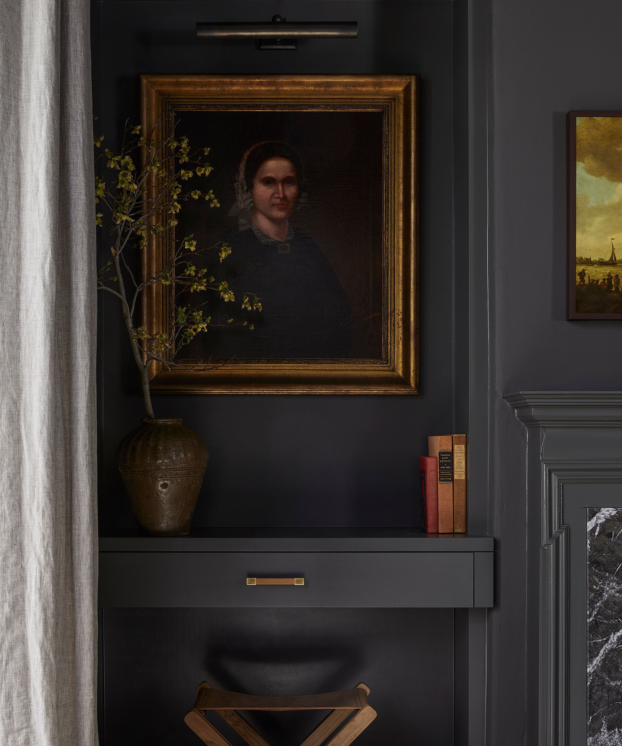 Close-up detail of a dark gray wall with a classic portrait in a gold frame illuminated by an art light, above a floating desk/shelf