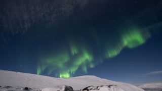 northern lights appear as dancing ribbons of green in the sky above a snowy mountain top. 