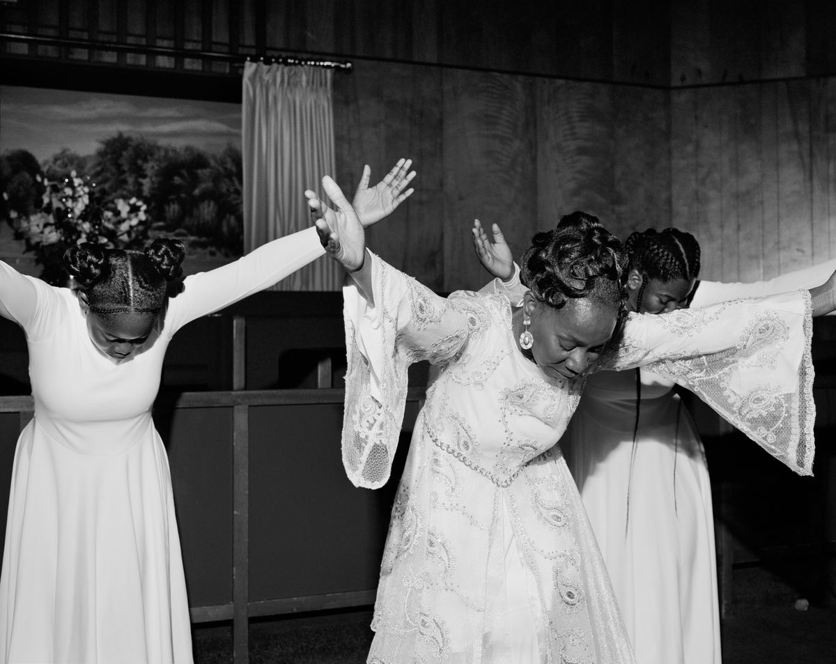 Black and white photograph of three women, wearing white dresses. They have their heads bent down with their arms outstretched.