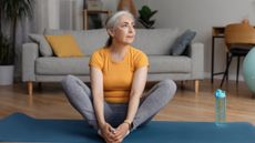 Woman sits on an exercise mat in a living room with the soles of her feet together