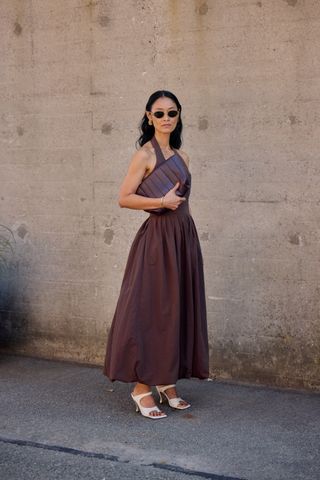 A woman wear oval-shaped sunglasses, a brown maxi dress, white heels, and a brown oversized clutch bag.