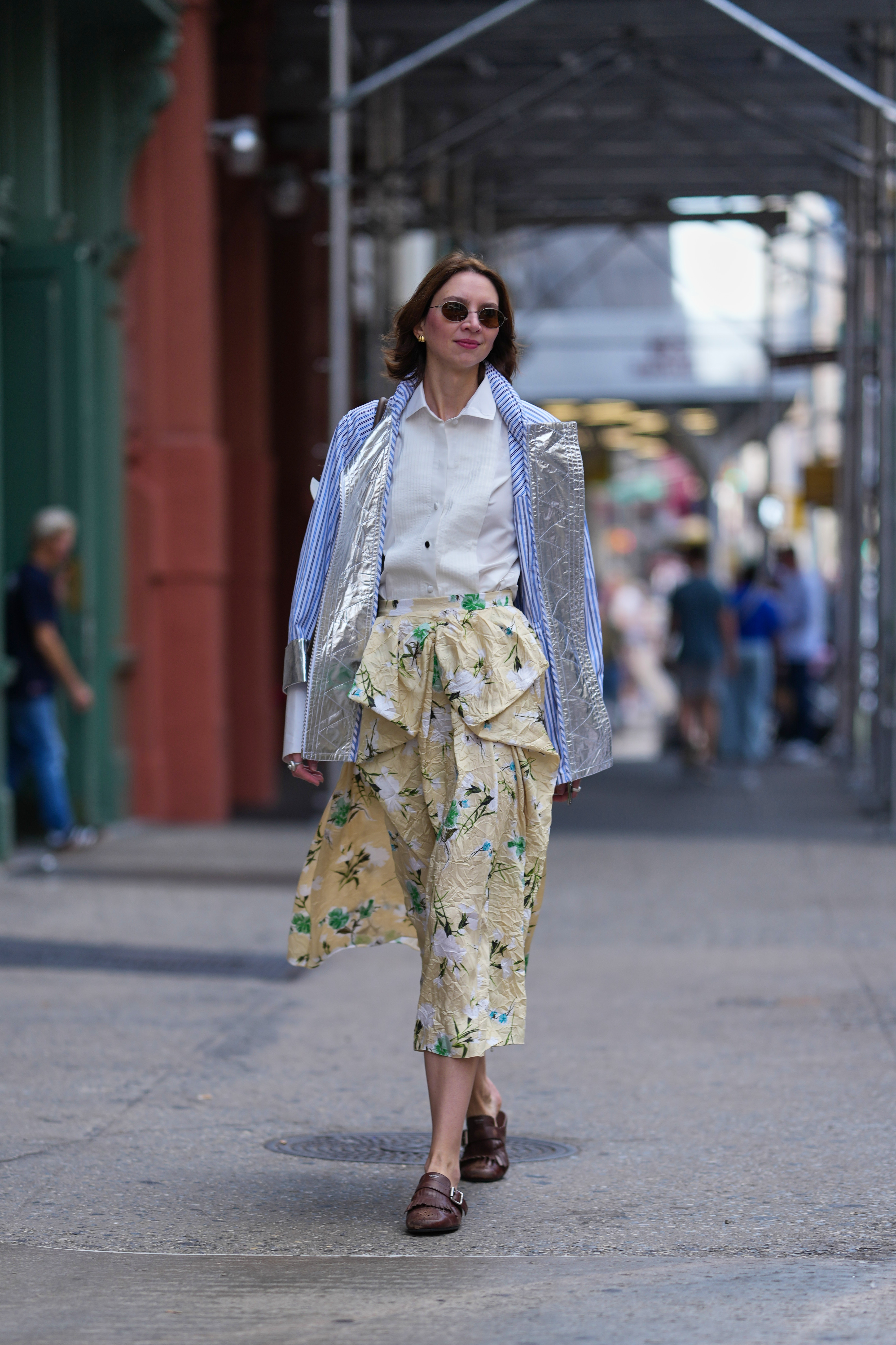 A guest wears shoulder-length brown hair in a loose bob, small oval sunglasses with tinted lenses and thin dark frames, small gold hoop earrings, a brown leather shoulder bag with a narrow strap, a blue-and-white striped blazer jacket with a silver metallic crinkled front panel worn over a white collared button-down shirt, a pale yellow crinkled midi skirt with green and white floral print and an oversized draped bow detail at the waist, brown leather loafers with buckle straps, outside Tibi, during New York Fashion Week, on September 13 2025 in New York, New York