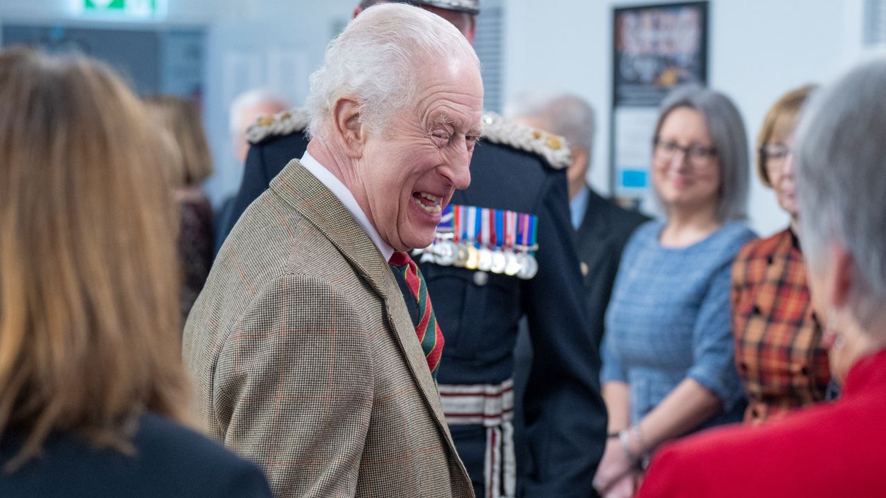 King Charles wearing a tan blazer and striped tie turning to the side and laughing surrounded by a group of people 