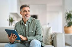 Happy mature man using digital tablet relaxing on couch looking away at home.