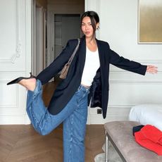 style influencer Mimi Nguyen poses with one leg up in a chic hotel in Monaco wearing a black blazer, white t-shirt, a taupe chain-strap bag, baggy barrel jeans, and black mule heels