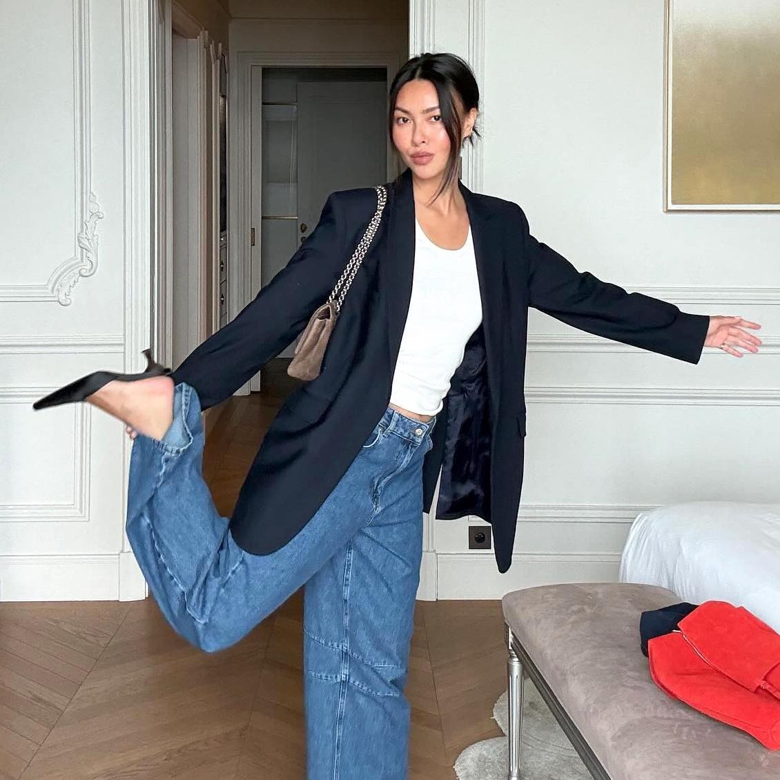 style influencer Mimi Nguyen poses with one leg up in a chic hotel in Monaco wearing a black blazer, white t-shirt, a taupe chain-strap bag, baggy barrel jeans, and black mule heels