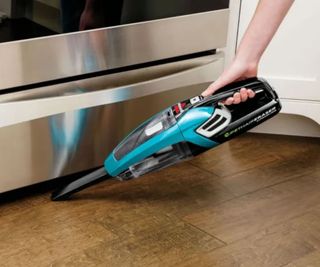 Bissell Handheld vacuum vacuuming under an oven drawer on a wooden surface