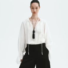 Model wears white top, black barrel-leg trousers, fringed necklace