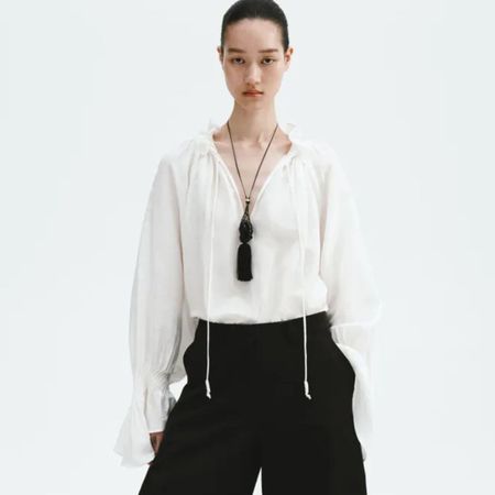 Model wears white top, black barrel-leg trousers, fringed necklace