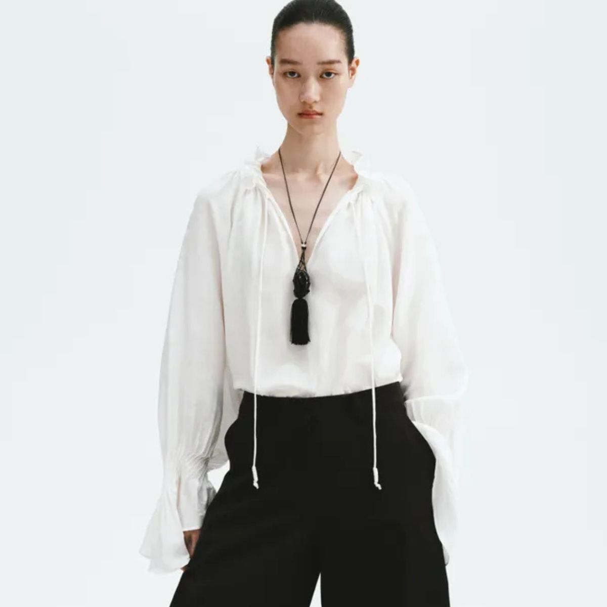 Model wears white top, black barrel-leg trousers, fringed necklace